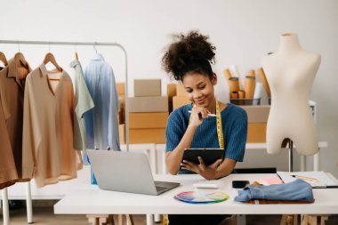 Gülümseyen mutlu Afro-Amerikalı terzi kadın elbise odasında çalışıyor ve masada otururken dijital tablet kullanıyor. 