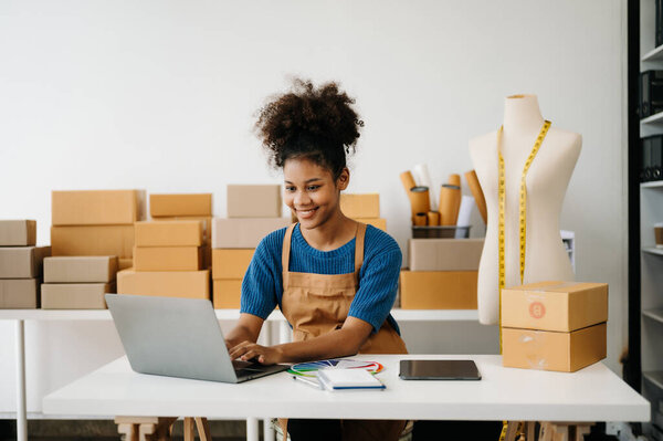 Афро-американская портная работает за столом с ноутбуком, цветной палитрой и цифровым планшетом, доставка коробки и тело манекена на заднем плане 