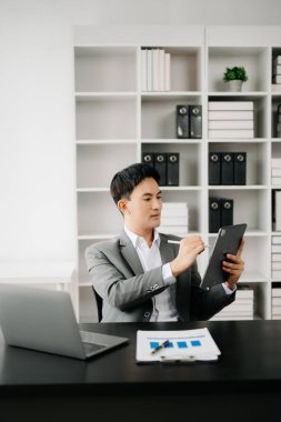 Modern ofiste çalışan genç Asyalı işadamı dizüstü bilgisayarı, tableti ve kâğıdı not alıyor.