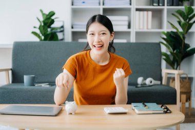 Asyalı kadınların eli, iş, finans, tasarruf ve gayrimenkul yatırım kavramı için masaya ahşap ve tablet koymak üzere tahtadan bir parayı domuz kumbarasına tasarruf ettiriyor. ev ofisinde.