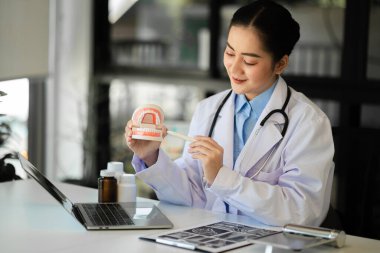 Diş örnekleriyle dolu bir masada oturan konsantre dişçi ve diş hekimliği kliniğinde tablet ve dizüstü bilgisayarla çalışıyor. Tıp doktoru çalışıyor.