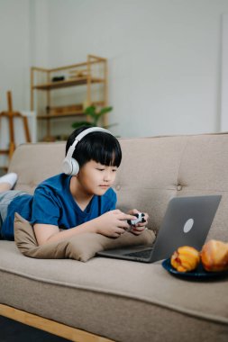 Genç Asyalı çocuk evde kulaklık takıp kanepede yatarken joystick ile video oyunu oynuyor. 