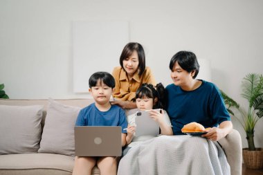 Asyalı ebeveynler ve çocuklar koltukta otururken birlikte cihaz kullanıyorlar. Teknoloji bağımlısı aile evde eğleniyor.