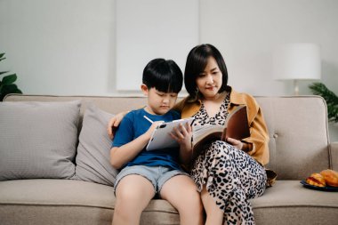 Çocuk tablet ve kadın kitap okurken Asyalı anne ve oğlu aile koltuğunda oturuyorlar.