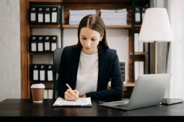 Kahve fincanı ve dizüstü bilgisayarla masada otururken siyah ceketli kadın notlar yazıyor. 