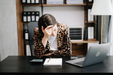 Ofis masasında çok çalışan bir iş kadını, leopar ceketi giyiyor, baş ağrısı çekiyor. 