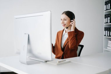 Kablosuz kulaklıkla çağrı merkezi operatörü. Müşteri ile konuşuyor. Kulaklıklı kadın, bilgisayar monitörüyle masada çalışırken mikrofon danışmanlığı yapıyor. 