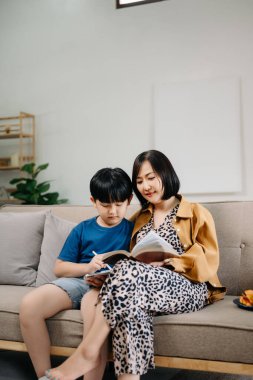 Mutlu Asyalı anne ve oğlu aile kanepesinde oturup birlikte kitap okuyorlar.