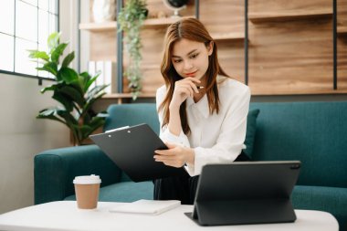 Genç ve güzel bir kadın çalışma yerinde otururken dizüstü bilgisayar ve tablet kullanıyor. Oturma odasında, ofisteki kanepede.