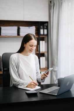 Kafkasyalı kadın dizüstü bilgisayarla masada oturuyor ve cep telefonu kullanan kağıt defterli kadına notlar yazıyor. 