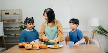 Asyalı bir aile mutfakta yemek yapıyor. Masanın yanında duran gülümseyen anne ve çocukların portresi..