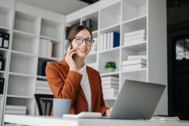 Ofis masasında laptopuyla cep telefonuyla konuşan beyaz bir kadın. 
