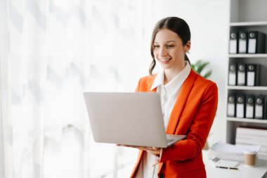 İş kadını, ofiste dikilirken dizüstü bilgisayar kullanıyor ve gülümsüyor ve kırmızı ceket giyiyor. 