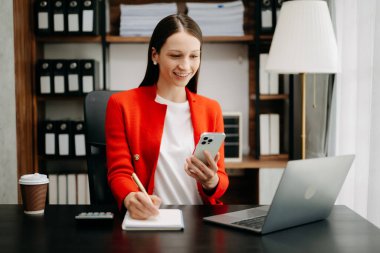 Beyaz iş kadını kırmızı ceket giyiyor, ofis masasında dizüstü bilgisayarla çalışıyor ve cep telefonu kullanıyor.,