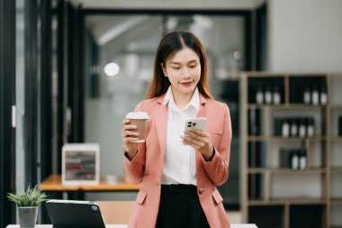 Gülümseyen, kahve içen ve akıllı telefon kullanan kendine güvenen Asyalı kadın modern ofis.