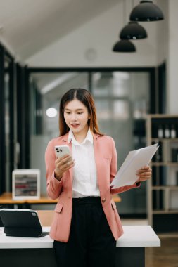 Modern ofiste elinde akıllı bir telefonla gülümseyen kendine güvenen Asyalı kadın.