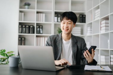 Genç işadamı ofiste dizüstü bilgisayar ve akıllı telefonla çalışıyor.