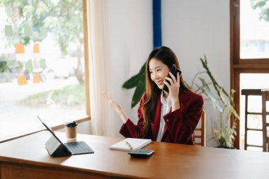 Asyalı iş kadını banka ofisinde dijital tabletle çalışıyor, gülümsüyor ve telefonla konuşuyor. 
