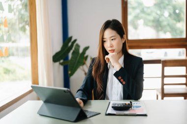 Asyalı iş kadını banka ofisinde dijital tabletle çalışıyor, elleri çenesinde düşünüyor.