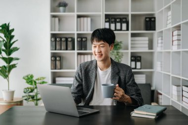 Genç Asyalı iş adamı modern ofiste dizüstü bilgisayar kullanıyor. Gülümseyen kendine güvenen beyaz yakalı işçi