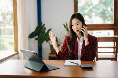 Asyalı iş kadını banka ofisinde dijital tabletle çalışıyor, telefonla konuşuyor ve ekrana bakıyor. 