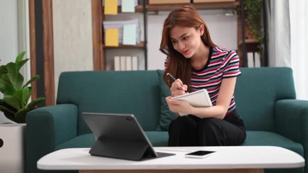 働く木のテーブルの現代オフィスに座っている間デジタル タブレットが付いているノートを作る若い美しい女性 — ストック動画