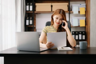 Asyalı iş kadını telefonda konuşuyor ve ofisteki bilgisayarı kullanıyor.