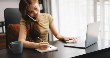 İş kadını eli dizüstü bilgisayarla çalışıyor ve not defterine notlar alıyor.