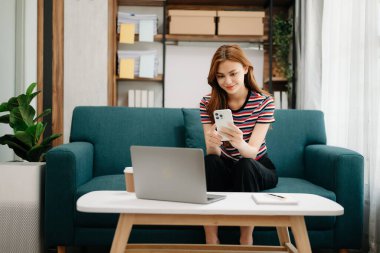 Genç ve güzel bir kadın iş yerinde otururken dizüstü bilgisayar ve telefon kullanıyor. Oturma odasında, ofisteki kanepede.