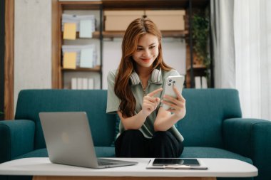 Kız öğrenci, Asyalı kız kütüphanesi koltukta oturuyor. Oturma odasında dizüstü bilgisayar ve telefon kullanıyor.