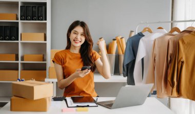 Moda blogcusu kavramı, giysi satan genç Asyalı kadın. Akıllı telefon ve dizüstü bilgisayar kullanarak küçük işletme KOBİ 'si kurun, ofiste satın alın ve kontrol edin