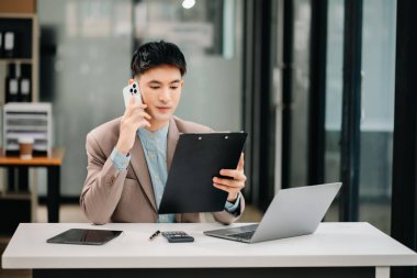 Genç Asyalı işadamı modern ofisindeki iş yerinde cep telefonuyla konuşuyor.