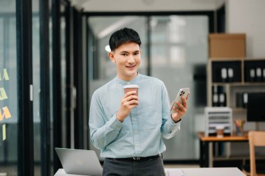 Genç ve çekici Asyalı işadamı kahve içiyor ve modern ofiste akıllı telefon kullanıyor.