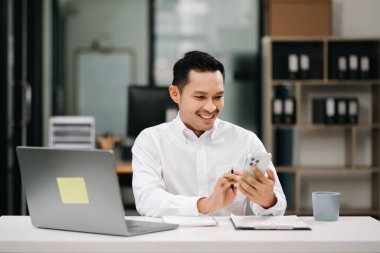Modern ofiste bilgisayar, akıllı telefon ve belgelerle çalışan gülümseyen Asyalı işadamı