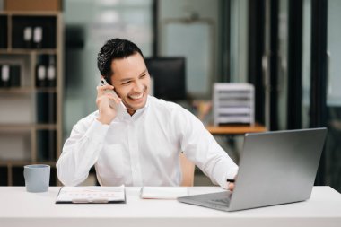 Modern ofiste bilgisayar ve belgelerle çalışan gülümseyen Asyalı işadamı cep telefonuyla konuşuyor.
