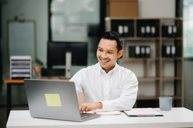 Modern ofiste bilgisayarla ve belgelerle çalışan gülümseyen Asyalı işadamı