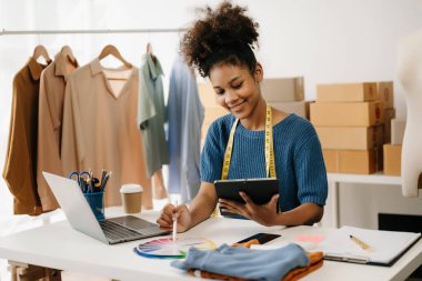 Koyu tenli kadın terzi tablet bilgisayarla çalışıyor. Stüdyodaki kıvırcık esmer moda tasarımcısı