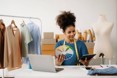 Koyu tenli kadın terzi tablet bilgisayarla çalışıyor. Stüdyodaki kıvırcık esmer moda tasarımcısı