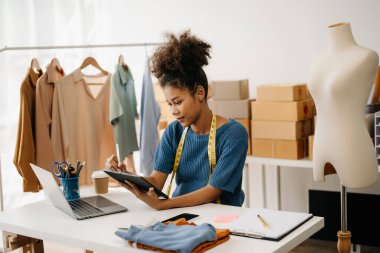 Koyu tenli kadın terzi tablet bilgisayarla çalışıyor. Stüdyodaki kıvırcık esmer moda tasarımcısı