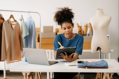 Koyu tenli kadın terzi tablet bilgisayarla çalışıyor. Stüdyodaki kıvırcık esmer moda tasarımcısı