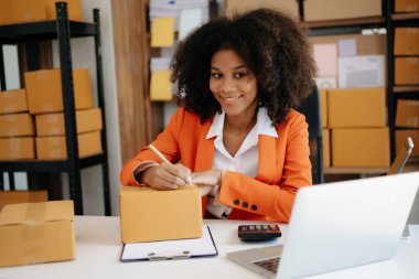 Küçük işletme KOBİ 'si kurun. Girişimci Afrikalı kadın internetten sipariş almak ve kontrol etmek için laptop kullanıyor. Genç bayan küçük işletme sahibi bunları müşterilere göndermek için paket paketliyor.