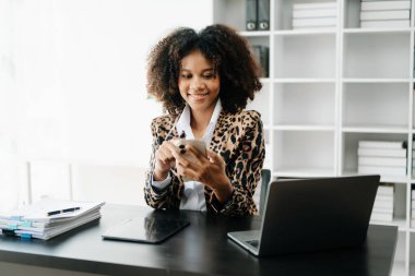 Afro saçlı genç Afrikalı kadın, ofiste çalışırken leopar ceketi giyiyor ve cep telefonu kullanıyor. 