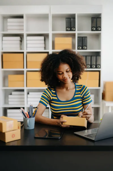 Stock image Startup small business SME. Entrepreneur African woman receiving and checking online orders. Young female small business owner packing parcels to ship them to customers