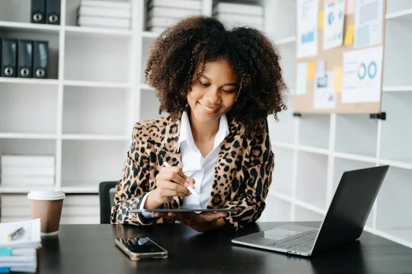 Afro saçlı genç Afrikalı kadın, ofiste çalışırken leopar ceketi giyiyor ve masada hesap makinesi ve dizüstü bilgisayarla tablet kullanıyor. 