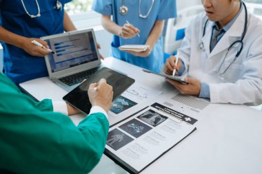 Tıbbi ekip, beyaz laboratuvar önlüklü ve ameliyat önlüklü doktorlarla toplantı yapıyor. Masada oturuyorlar ve tıp endüstrisinde dijital tablet ve dizüstü bilgisayar kullanıyorlar.