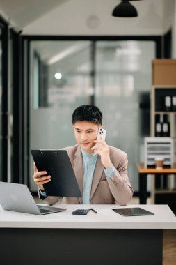 Gülümseyen Asyalı işadamı telefonda konuşuyor ve ofiste dizüstü bilgisayar kullanıyor.