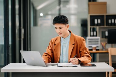Genç Asyalı iş adamı modern ofiste dizüstü bilgisayar kullanıyor. Gülümseyen kendine güvenen beyaz yakalı işçi