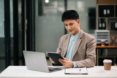 Genç Asyalı iş adamı modern ofiste dizüstü bilgisayar kullanıyor. Gülümseyen kendine güvenen beyaz yakalı işçi
