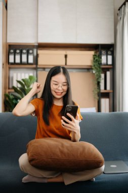 Çekici, mutlu Asyalı öğrenci üniversite kütüphanesinde okuyor, kanepede oturuyor, akıllı telefon kullanıyor.