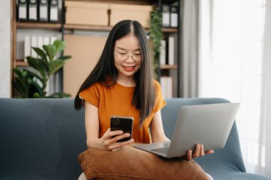  Asyalı kız kütüphanesi kanepede dizüstü bilgisayar ve tabletle internette arama yapıyor. 